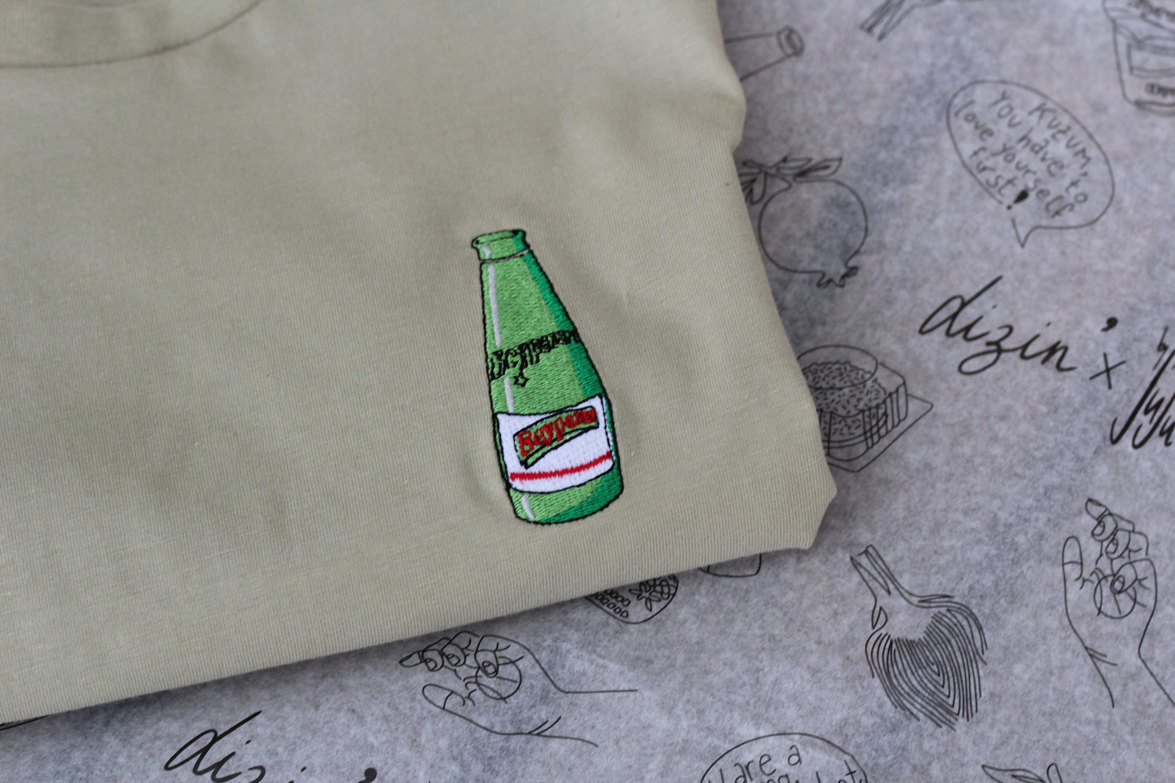Ein gefaltetes T-Shirt in sage Green mit einer gestickten Beypazari gazoz Flasche auf der Brust.